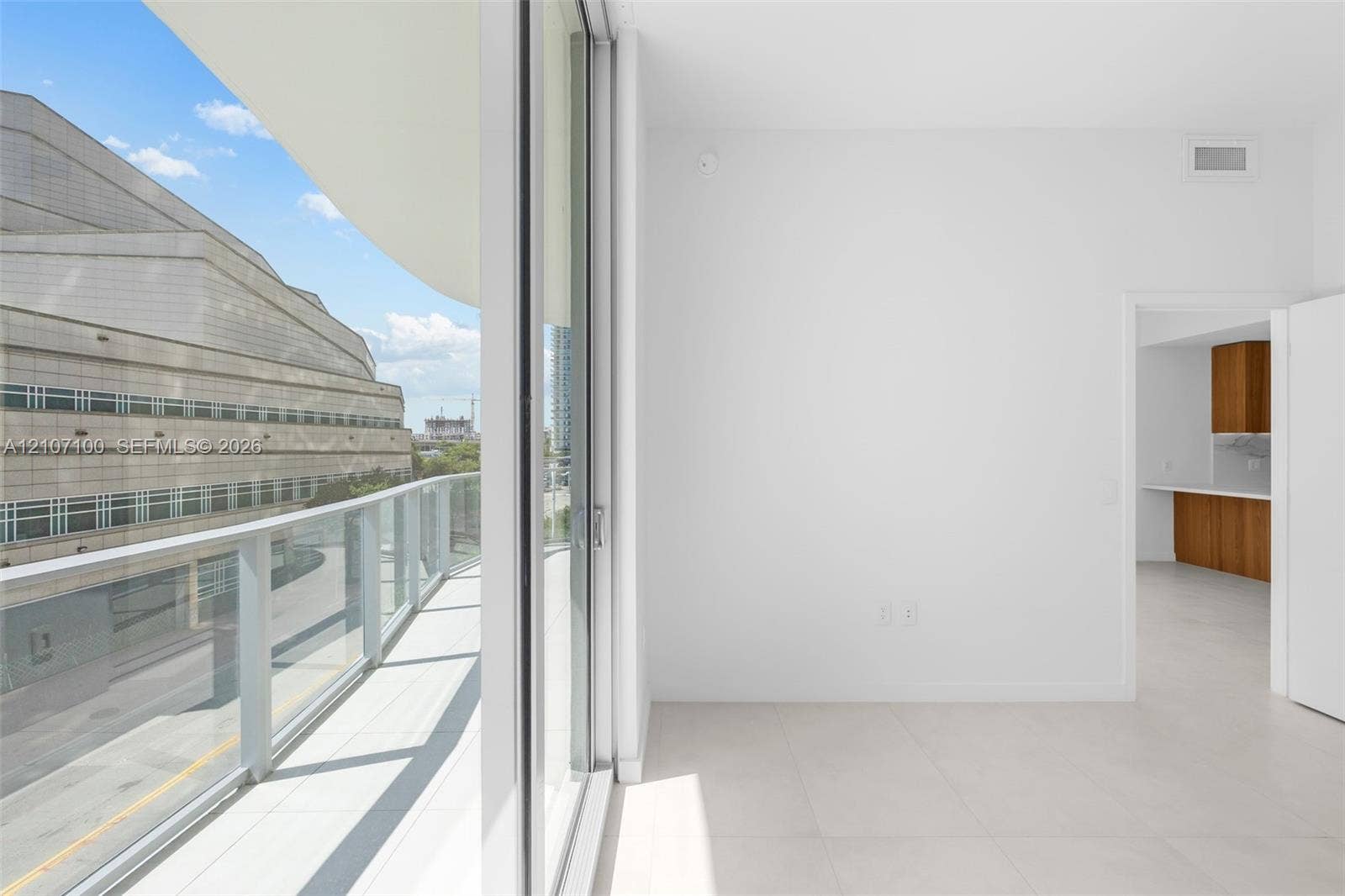 Floor-to-ceiling sliding glass doors opening to a covered balcony with a glass panel railing, paired with large-format tile flooring and wood-finish cabinetry