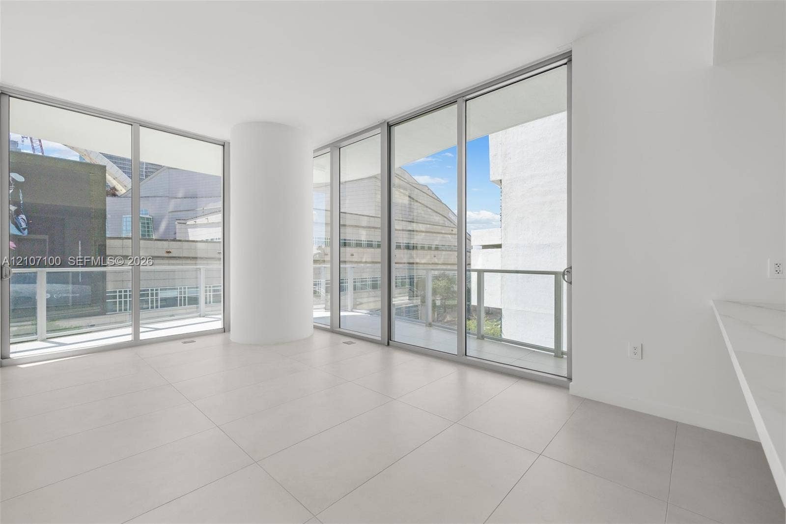 Floor-to-ceiling glass walls, expansive balcony access, neutral large-format tile flooring, and integrated white stone-finish countertop