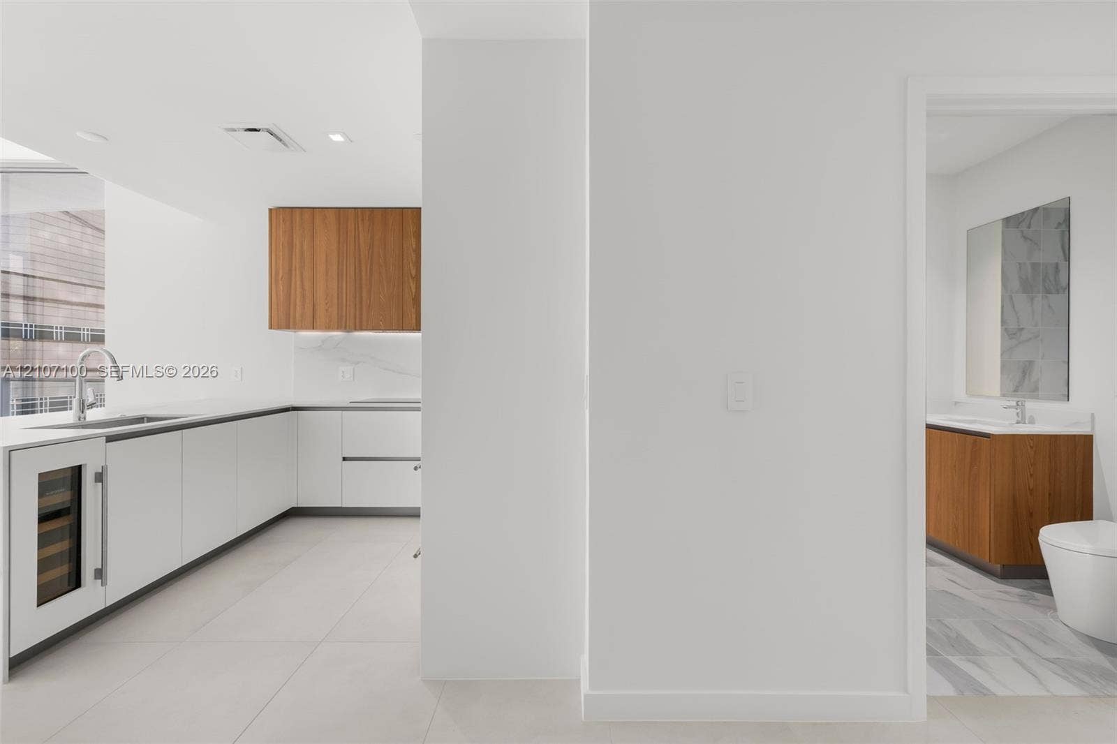 Kitchen featuring white handle-less cabinetry, wood-finish upper cabinets, integrated wine refrigerator, and stone-finish backsplash, with a restroom highlighting a floating wood-finish vanity and stone-tile flooring