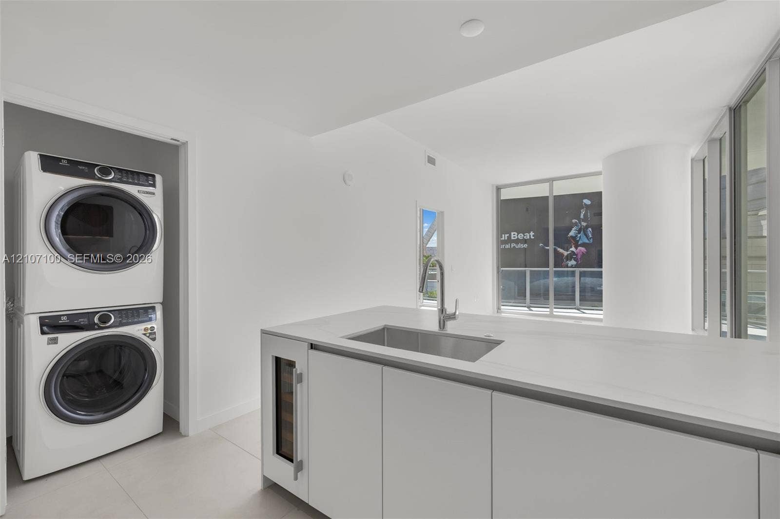 Kitchen island featuring a stone-surface countertop, undermount stainless steel sink, gooseneck faucet, and built-in wine cooler, accompanied by a stacked laundry appliance configuration