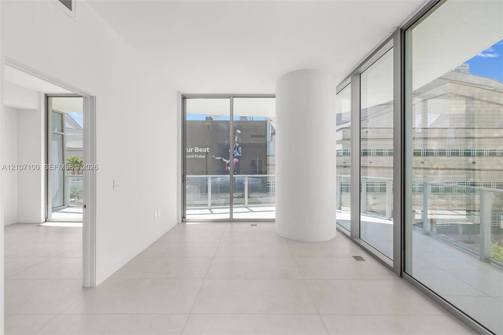 Floor-to-ceiling glass sliding doors, large-format tile flooring, a prominent structural column, and access to a perimeter balcony