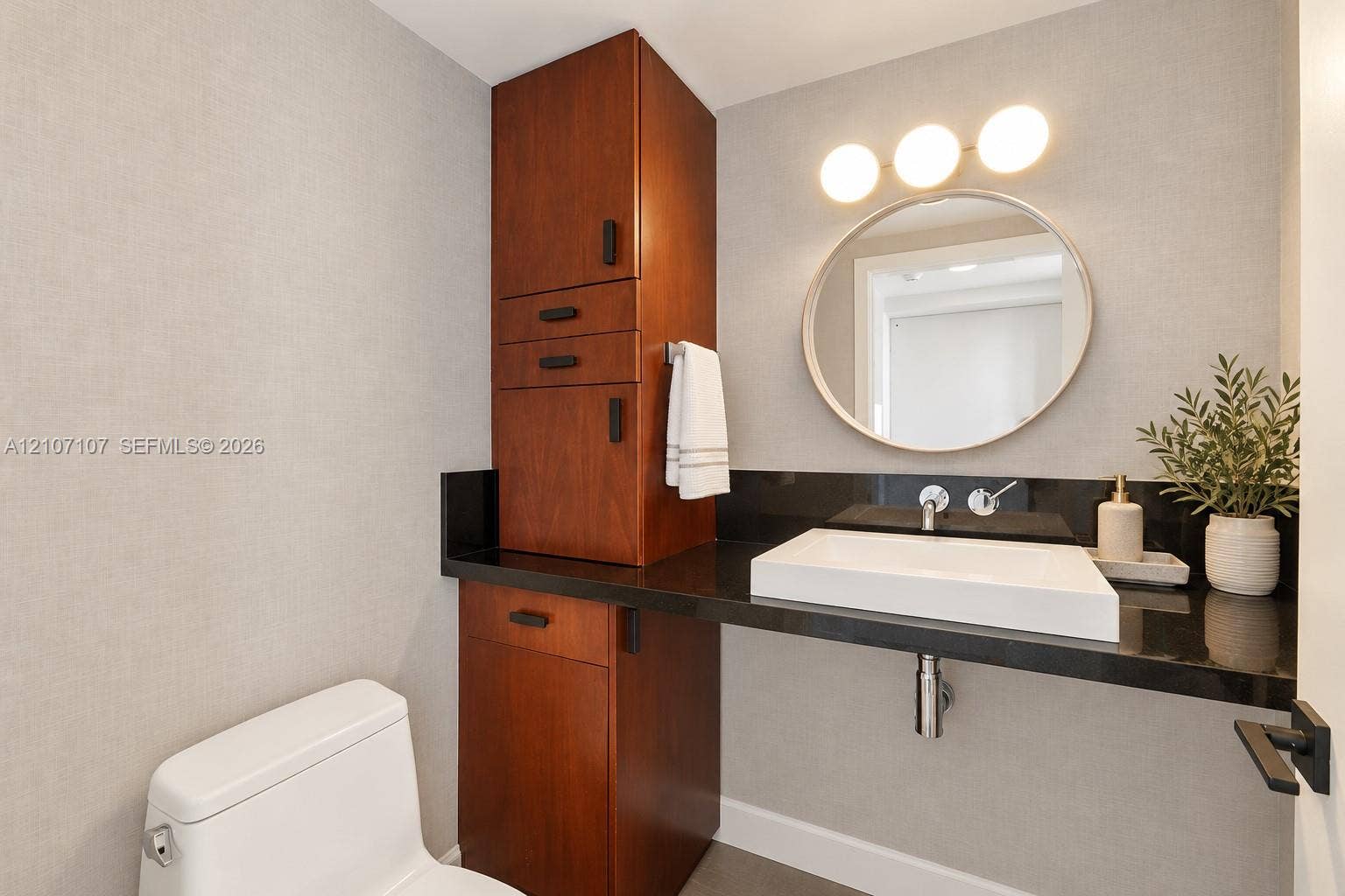 Built-in wood-finish cabinetry, black countertop, vessel sink, wall-mounted faucet, and circular vanity mirror with three-bulb integrated lighting