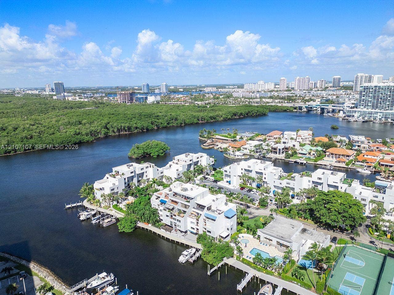 Aerial perspective of a waterfront residential complex featuring boat slips, a swimming pool, a tennis court, and dense coastal vegetation