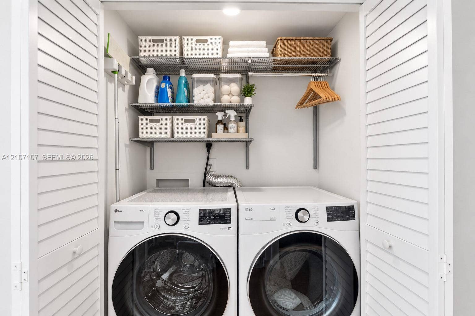 Laundry closet featuring dual white front-load appliances, louvered bi-fold doors, and integrated wire shelving for overhead storage