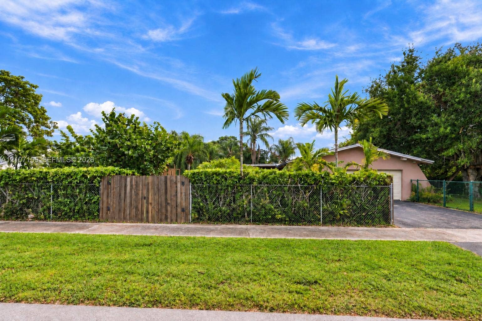 Exterior grounds featuring a metal chain-link perimeter fence, a solid wood privacy gate, mature tropical landscaping, and an asphalt driveway