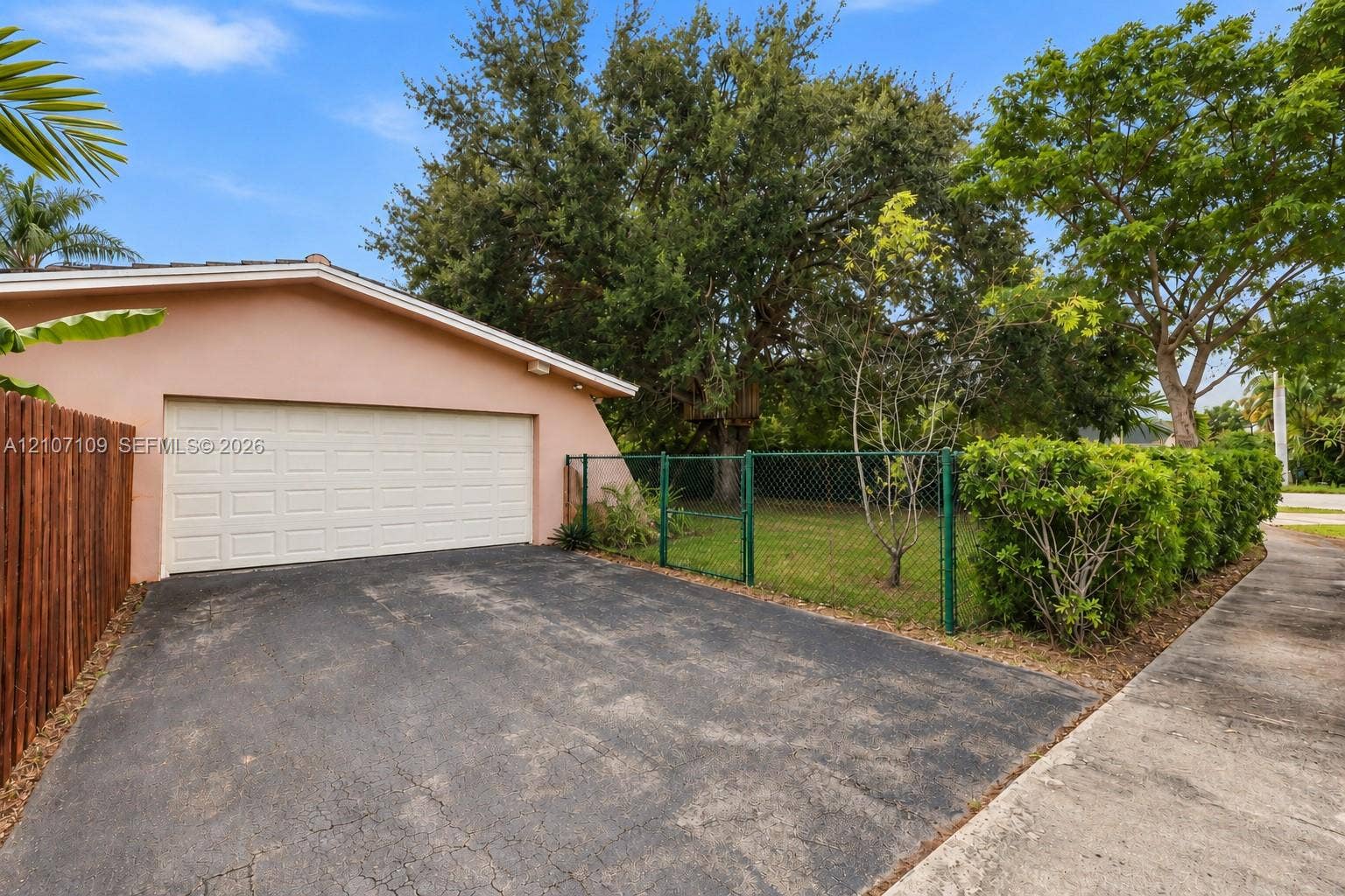 Attached garage with a wide sectional door, paved driveway, chain-link perimeter fencing, and mature trees on a grass-covered lot