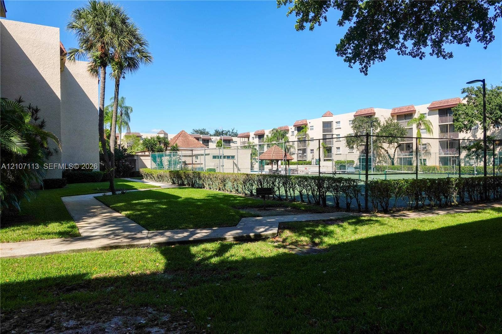 10451 W Broward Boulevard, #102, Plantation, Plantation, FL 33324 Recreational area featuring a fenced tennis court, manicured lawn, concrete walkways, and mature palm trees