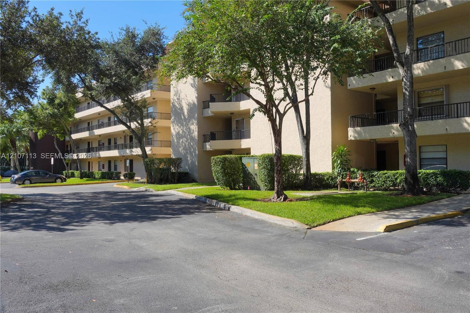 10451 W Broward Boulevard, #102, Plantation, Plantation, FL 33324 Multi-story building exterior featuring private balconies with metal railings, stucco facade, and paved parking area surrounded by mature landscaping