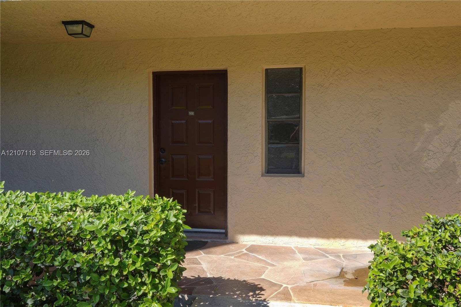 10451 W Broward Boulevard, #102, Plantation, Plantation, FL 33324 Entrance featuring a six-panel dark wood door, stucco exterior finish, flagstone paving, and flush-mount exterior lighting