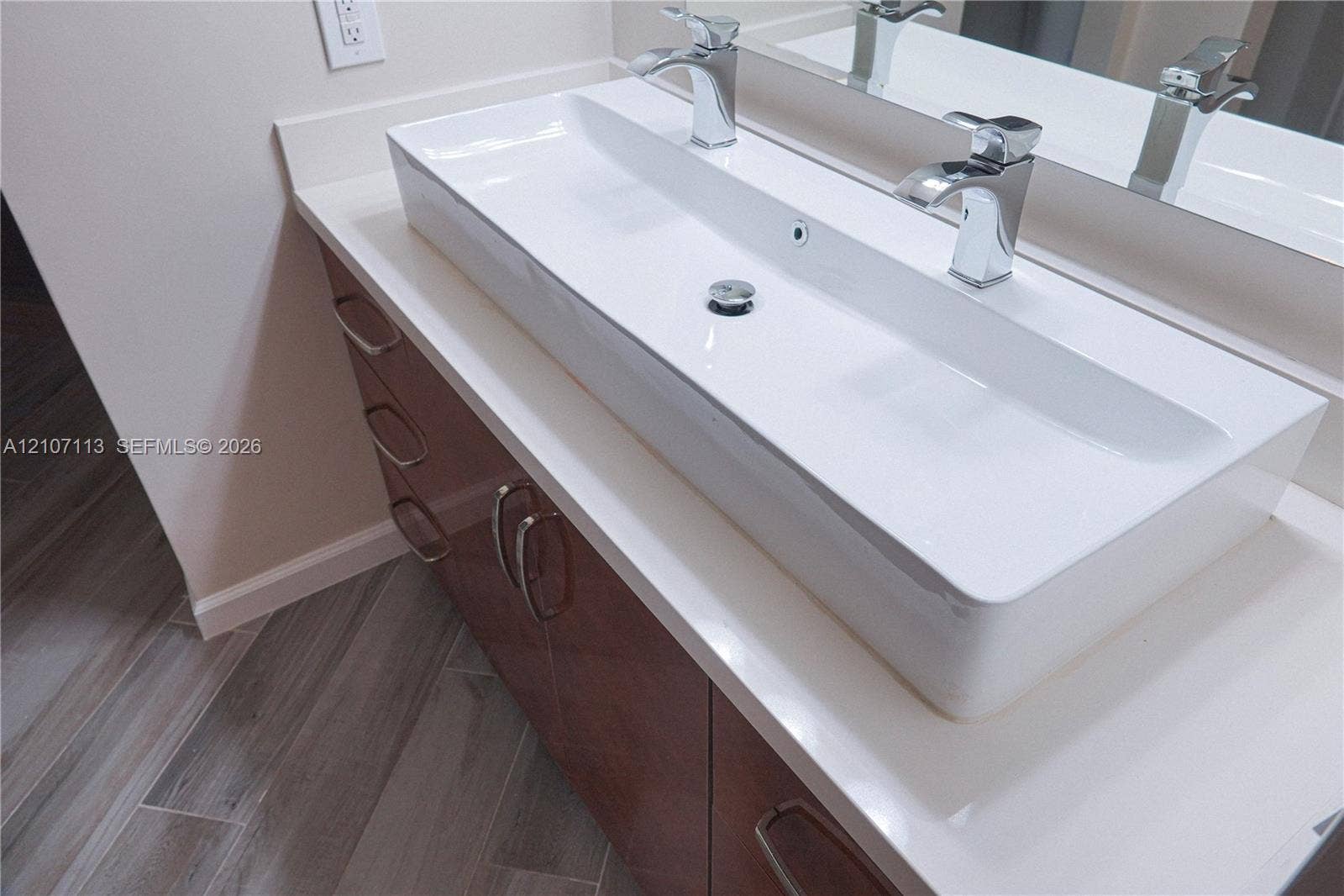 10451 W Broward Boulevard, #102, Plantation, Plantation, FL 33324 Bathroom vanity featuring a long, rectangular integrated sink, dual chrome faucets, solid-surface countertop, dark-toned cabinetry with metallic hardware, and wood-finish flooring