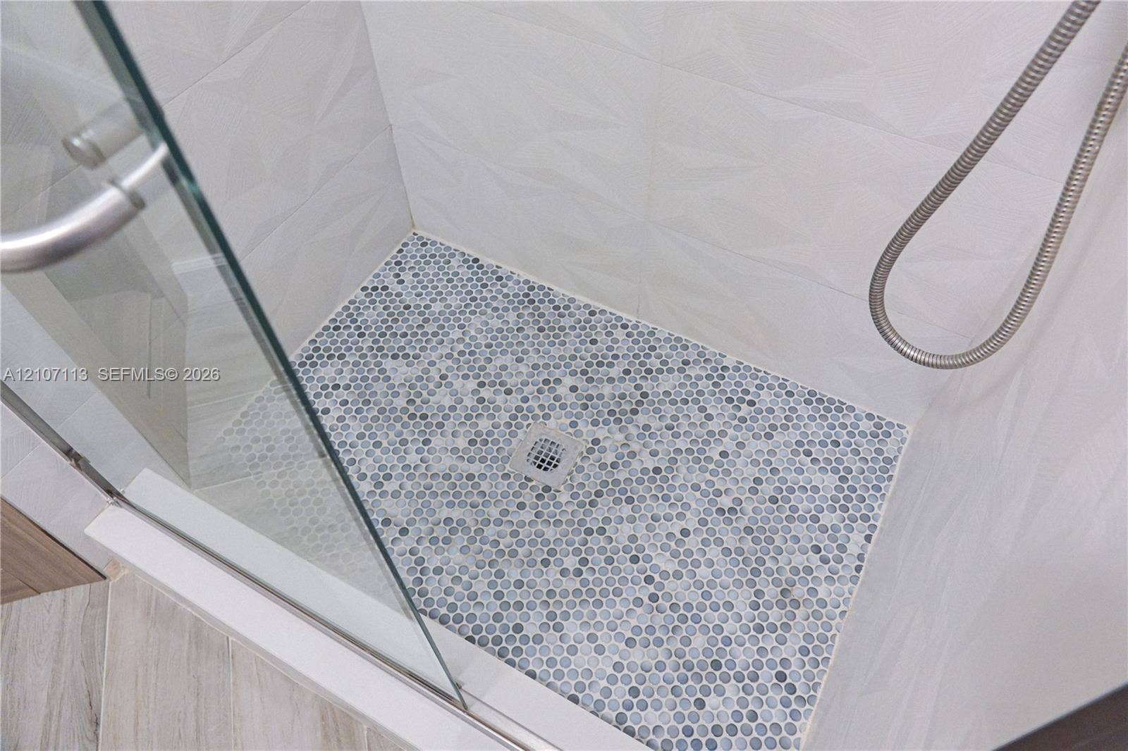 10451 W Broward Boulevard, #102, Plantation, Plantation, FL 33324 Walk-in shower featuring a frameless glass door, multi-faceted geometric wall tile, penny-round mosaic floor tiling, and a polished metal handheld shower fixture
