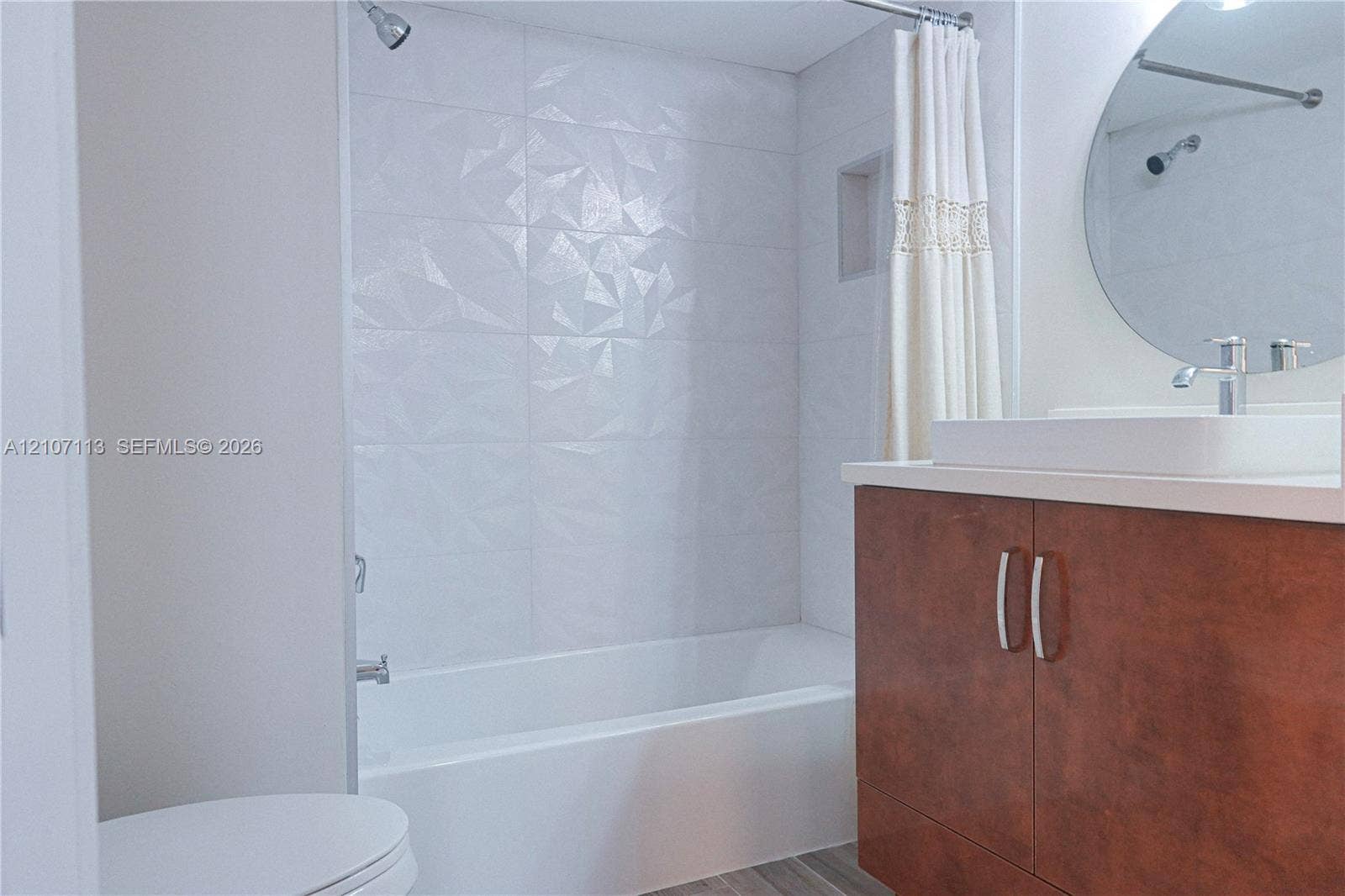 10451 W Broward Boulevard, #102, Plantation, Plantation, FL 33324 Bathroom featuring a wood-finish vanity with a rectangular basin, geometric textured tile shower surround, built-in shower niche, and a circular wall-mounted mirror