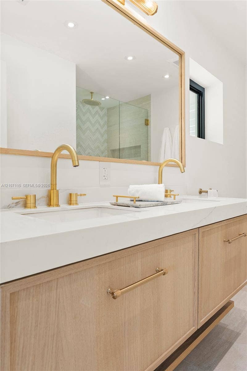 Floating vanity with light wood-finish cabinetry, solid surface countertop, dual undermount sinks, brass-toned hardware, and a framed mirror