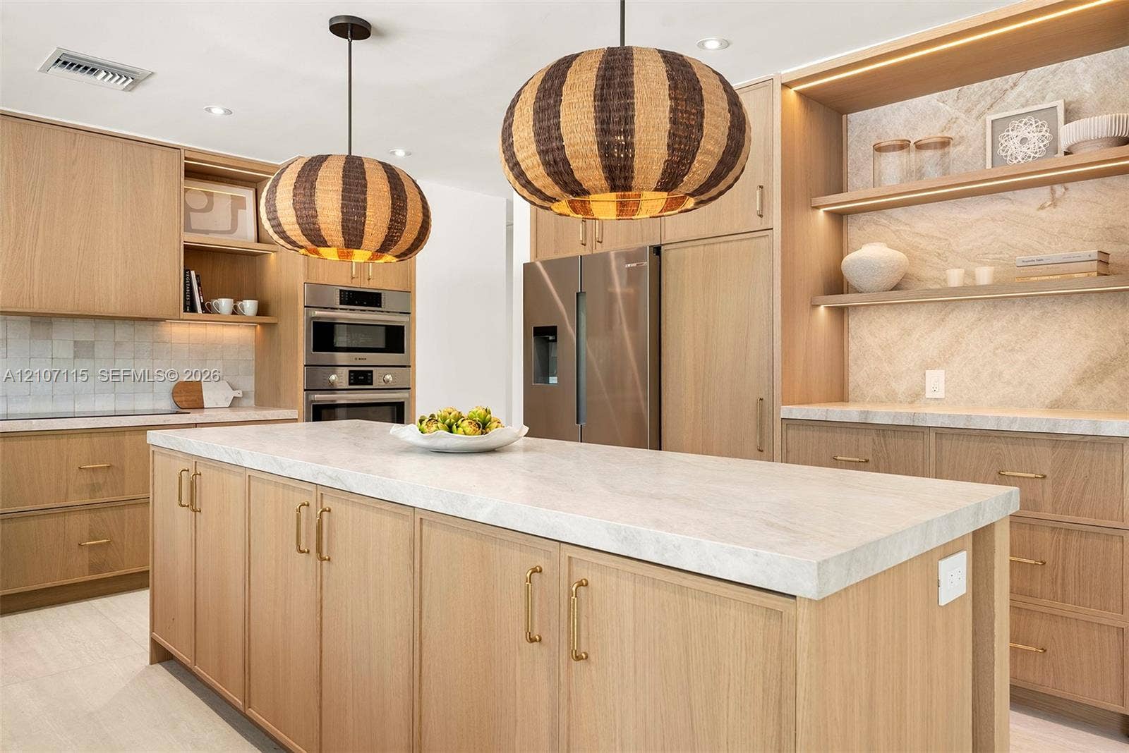 Kitchen featuring light wood-finish cabinetry, a waterfall-edge stone countertop island, built-in stainless steel appliances, decorative textured pendant lighting, and integrated shelf backlighting