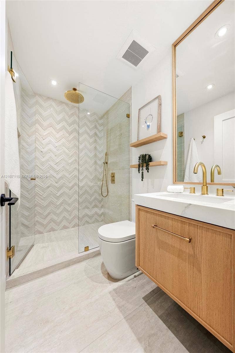 Wood-finish floating vanity with a white countertop and brass hardware, glass-enclosed walk-in shower with herringbone tile walls, and a large wall-mounted mirror with a wood frame