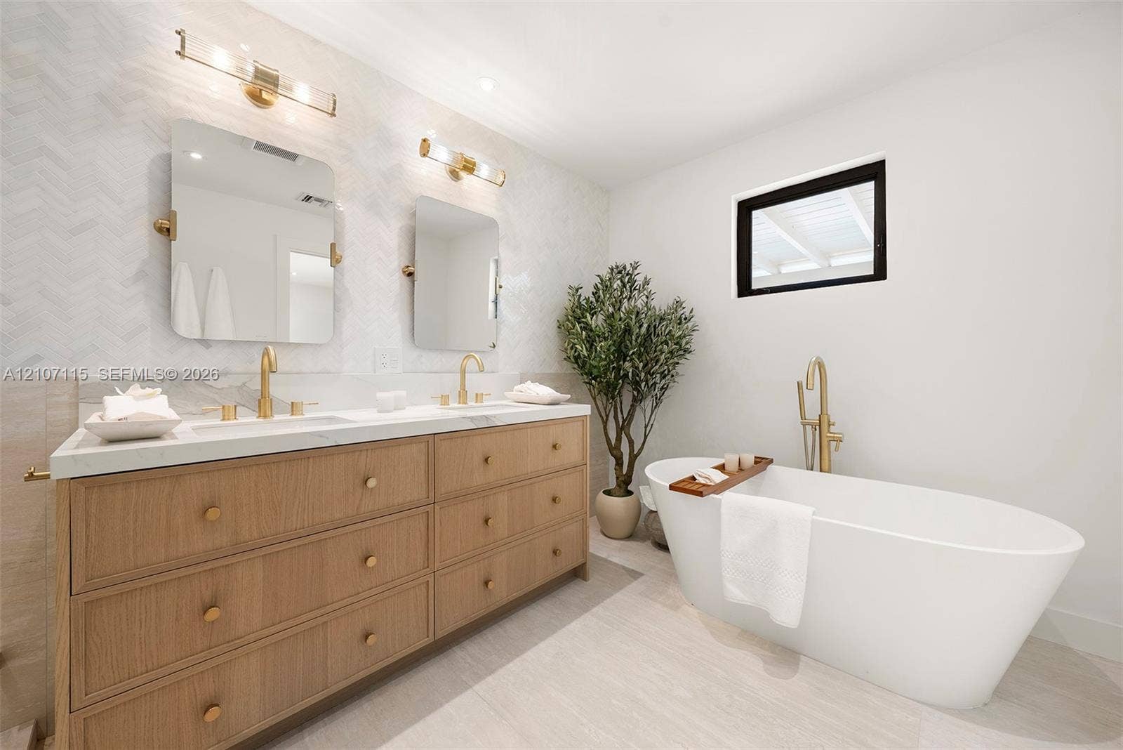 Double vanity with wood-finish cabinetry, quartz countertops, herringbone-patterned tile backsplash, polished brass fixtures, and a freestanding soaking tub