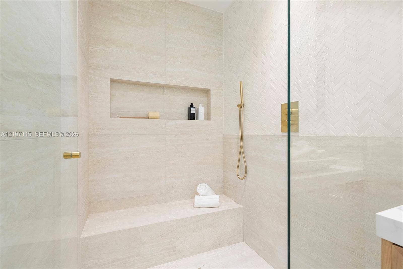 Walk-in shower featuring herringbone-patterned tile wall accents, built-in bench seating, recessed wall niche, and gold-finish hardware behind a glass enclosure