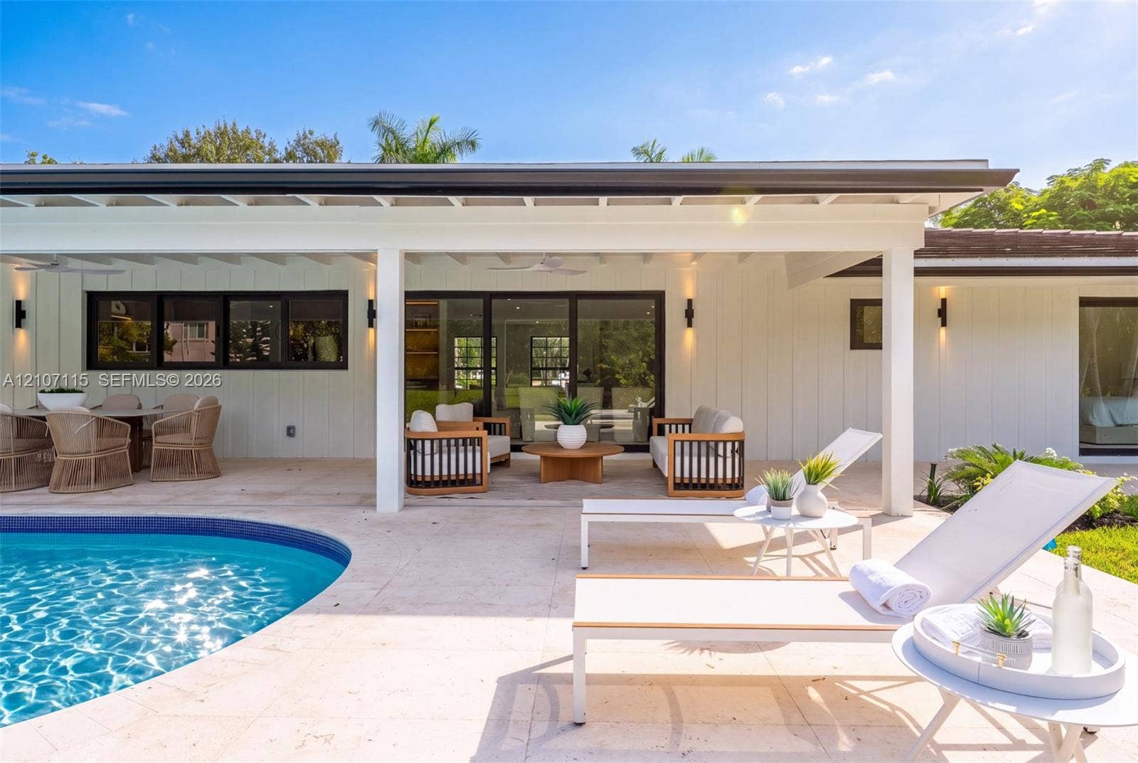 Covered outdoor patio featuring board-and-batten siding, recessed lighting, multi-panel sliding glass doors, and a stone-paver pool deck surrounding an in-ground swimming pool