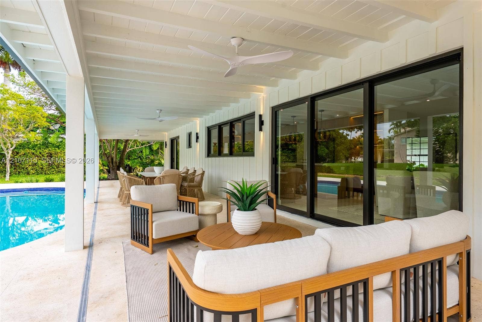 Covered outdoor patio featuring white tongue-and-groove ceiling, dual modern ceiling fans, sliding glass wall panels, and stone-finish flooring overlooking a private swimming pool