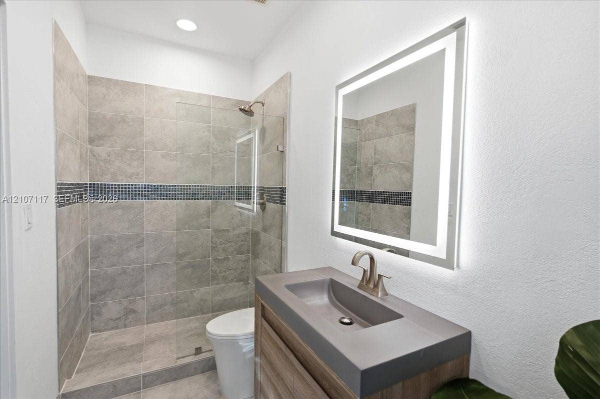 7863 NW 111th Court, Doral, FL 33178 Bathroom featuring a wood-finish vanity with a molded sink, LED-backlit mirror, walk-in shower with large-format tile and a glass partition, and a mosaic tile inlay