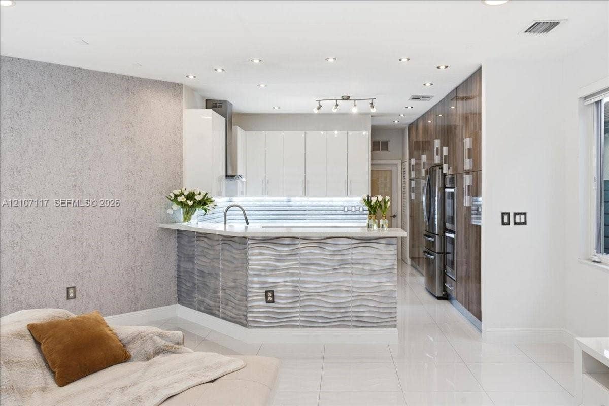 7863 NW 111th Court, Doral, FL 33178 Contemporary kitchen featuring high-gloss white upper cabinetry, full-height wood-finish pantry storage, a raised counter with a textured wave-pattern facade, integrated stainless steel appliances, and polished tile flooring