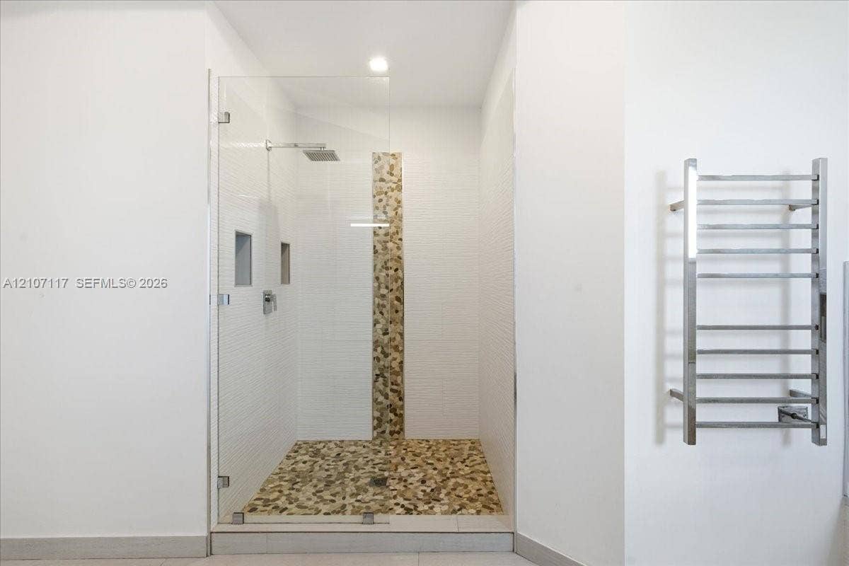 7863 NW 111th Court, Doral, FL 33178 Walk-in shower featuring a frameless glass door, stone-tile flooring and wall accent, dual recessed wall niches, rainfall showerhead, and a wall-mounted metal towel warmer