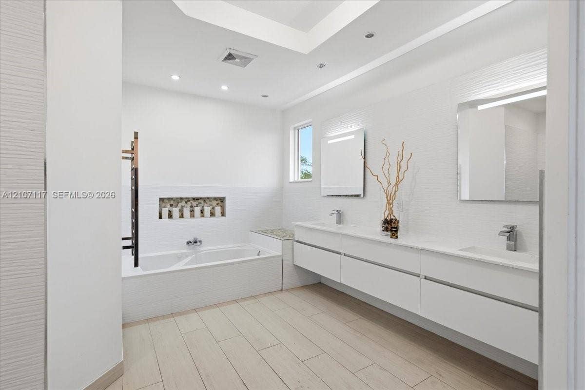 7863 NW 111th Court, Doral, FL 33178 Modern bathroom featuring a long floating dual-sink vanity with a solid-surface countertop, an inset tiled soaking tub with a decorative stone-accented recessed niche, wood-finish flooring, and wall-mounted mirrors with integrated lighting