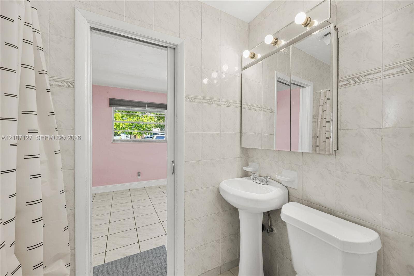 Full-height tile wall surround, white porcelain pedestal sink, mirrored medicine cabinet with integrated vanity lighting, and standard toilet