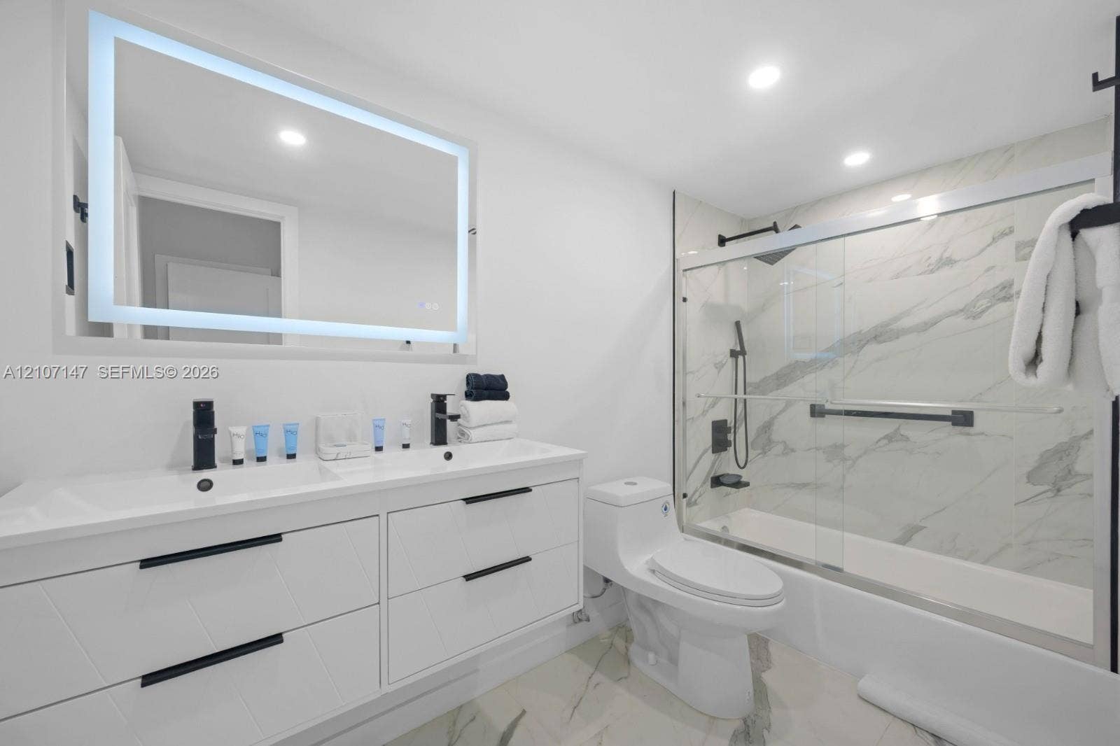 Modern bathroom featuring a double-sink vanity with matte black hardware, backlit LED mirror, recessed ceiling lighting, and a bathtub-shower combination enclosed by a glass door with large-format stone-patterned tile surround