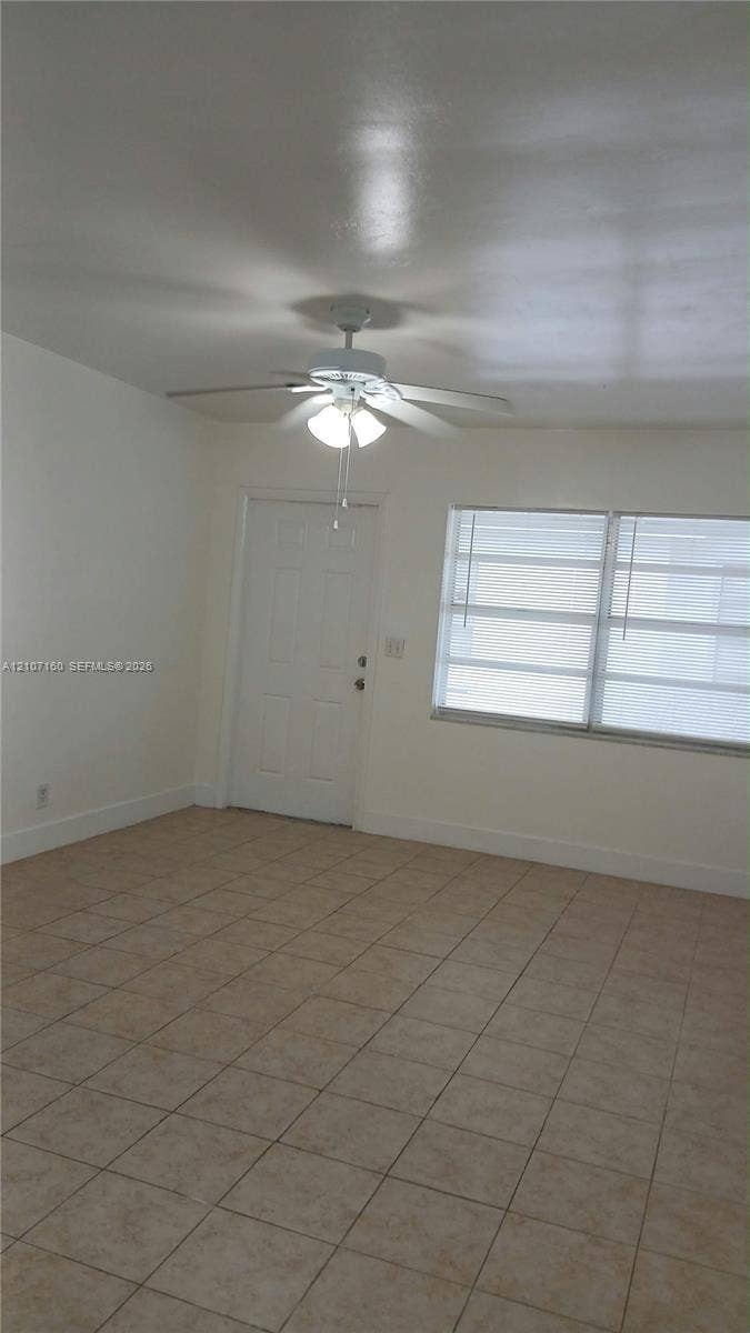 325 NE 2nd Street, #2, Hallandale Beach, Hallandale Beach, FL 33009 Interior space featuring neutral-toned tile flooring, multi-pane window with horizontal blinds, six-panel entry door, and ceiling-mounted light fixture with fan