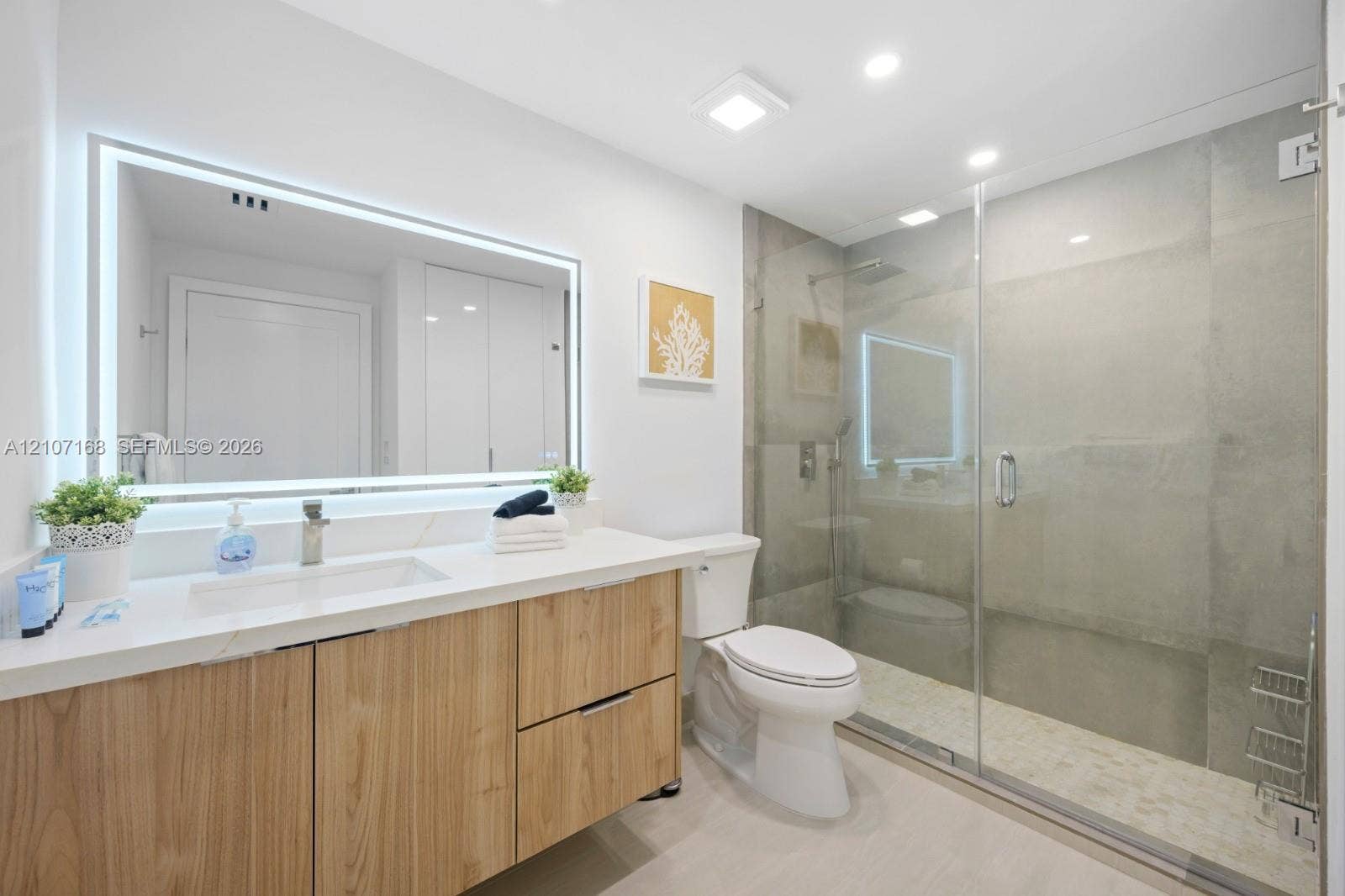 Bathroom featuring a wood-finish vanity with a stone countertop, an integrated rectangular sink, a frameless glass-enclosed shower, and a large backlit mirror