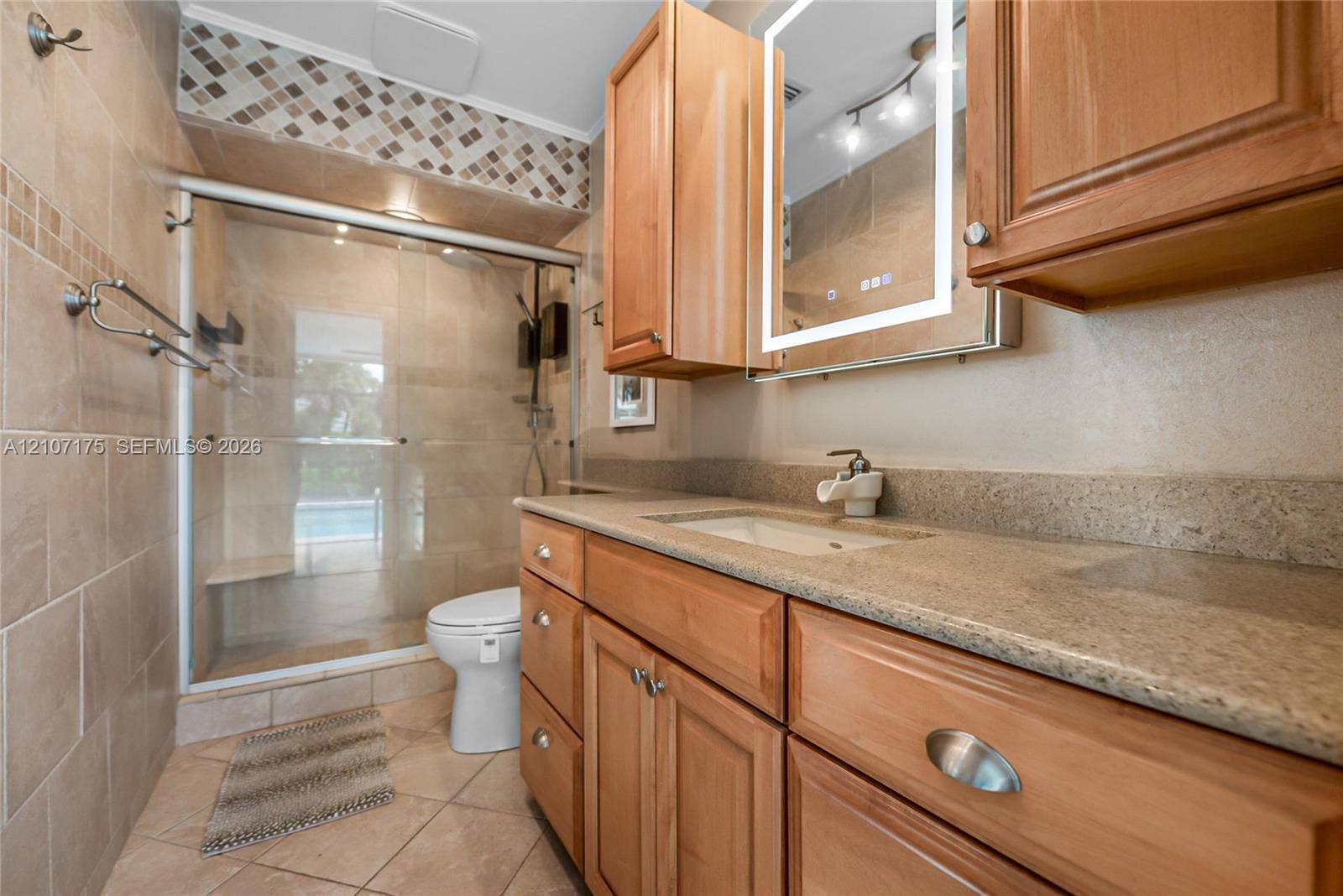 17550 SW 86th Avenue, Palmetto Bay, FL 33157 Bathroom featuring a wood-tone vanity with illuminated wall-mounted mirrored cabinet, walk-in shower with glass enclosure, and ceramic tile flooring