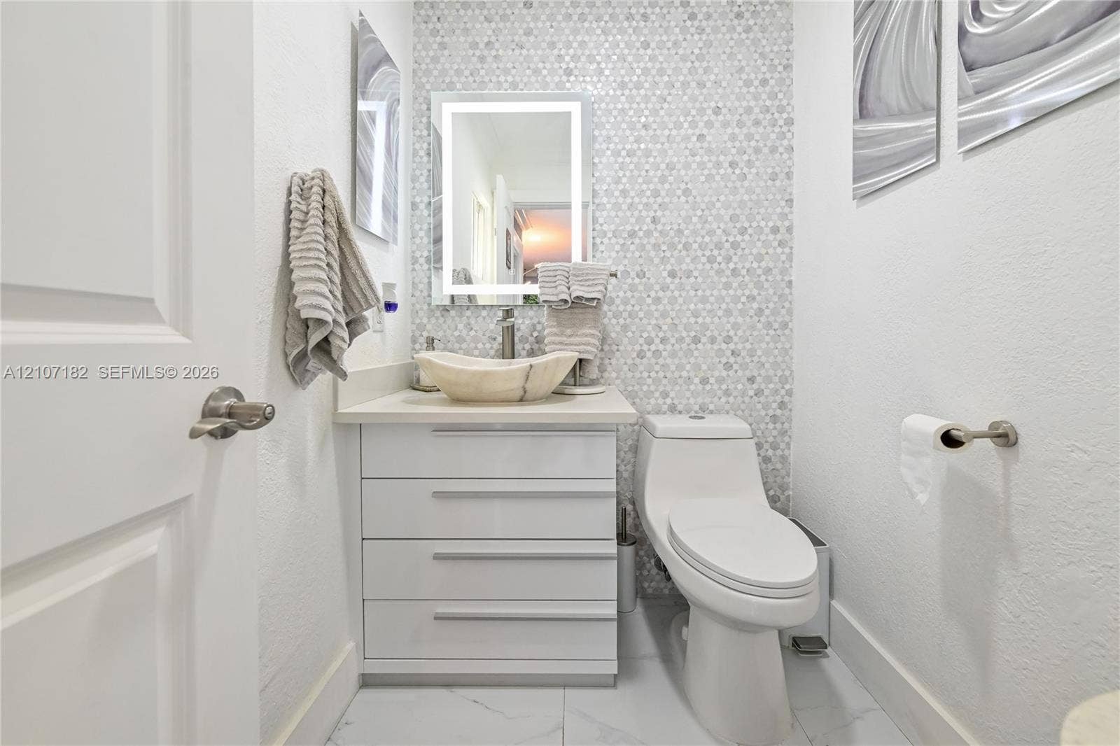 Bathroom featuring a stone vessel sink, white vanity with integrated cabinetry, mosaic tile wall accent, and illuminated vanity mirror
