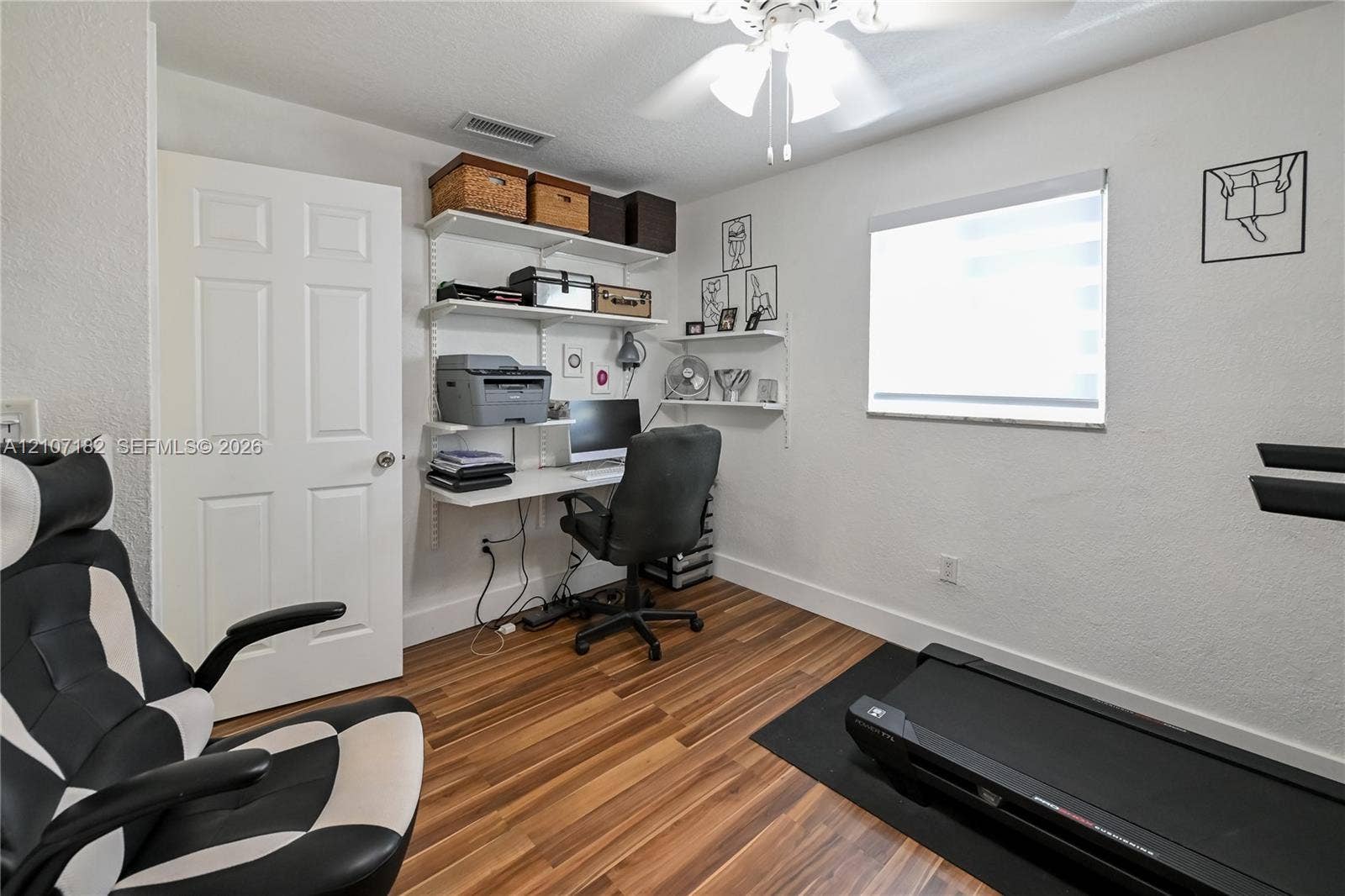 Wood-finish flooring, white six-panel interior door, wall-mounted adjustable shelving system, ceiling fan with integrated lighting, and single-pane window