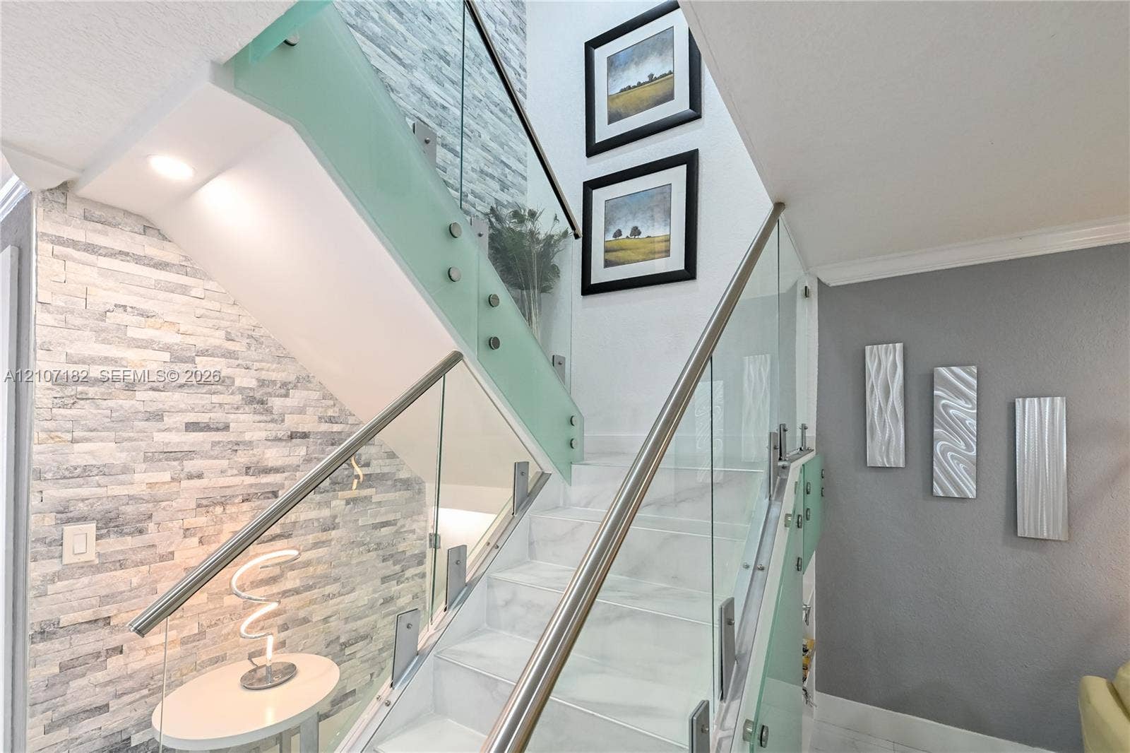 Staircase featuring white stone-finish treads, glass panel railing with stainless steel hardware, and an accent wall composed of stacked ledger stone