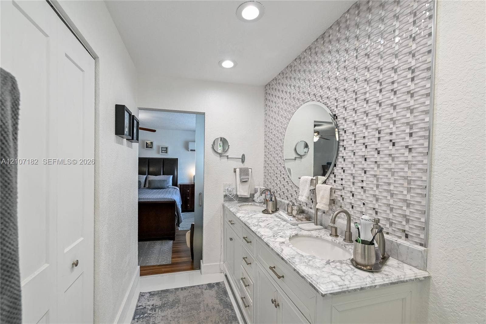 Dual-vanity cabinetry with stone-finish countertops, mosaic-tiled accent wall, circular wall-mounted mirror, and recessed lighting