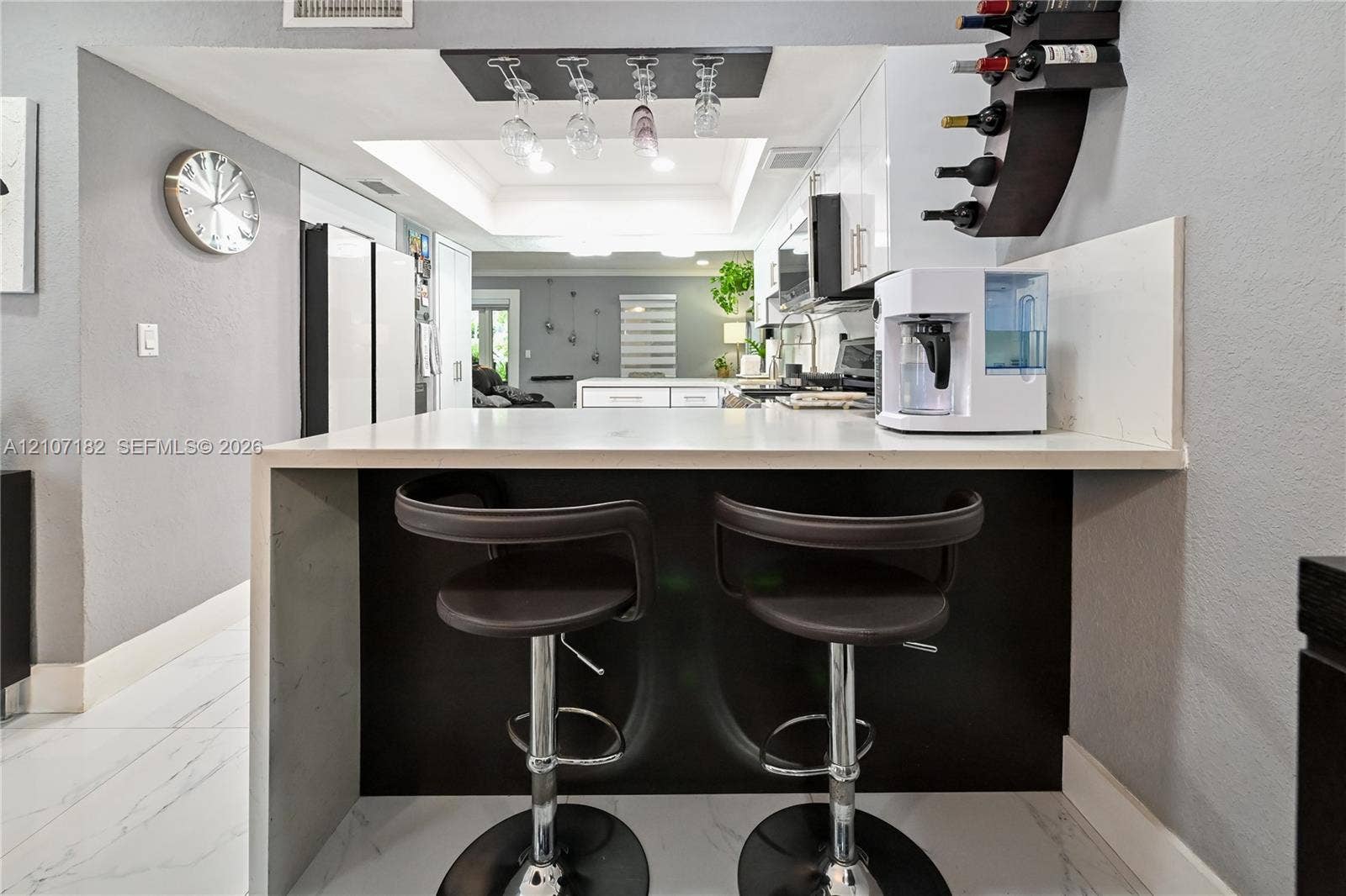 Waterfall peninsula featuring a solid-surface countertop and contrasting dark cabinetry, overhead wine glass rack, tray ceiling, and white floor tiling with veining