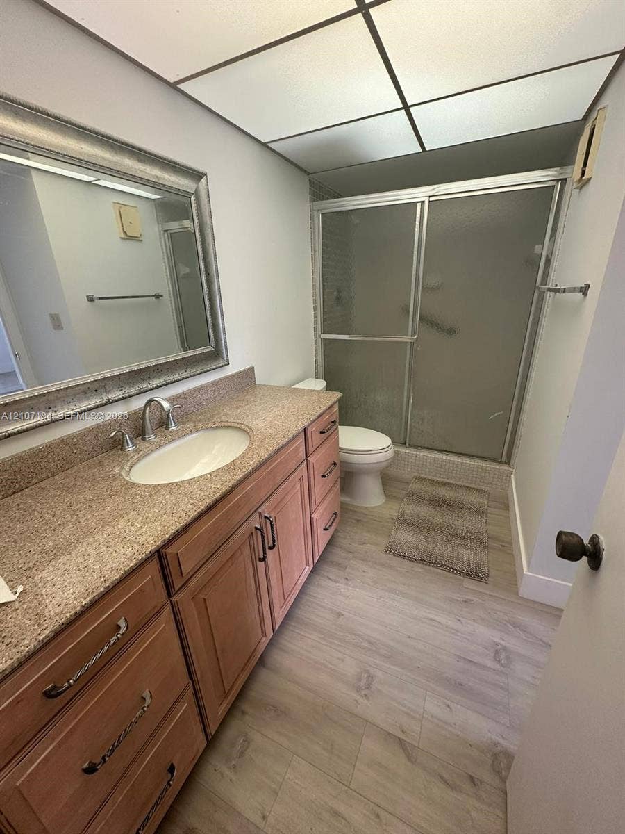 240 Lakeview Drive, #209, Weston, Weston, FL 33326 Bathroom featuring a wood-finish vanity with a stone-surfaced countertop, integrated oval basin, walk-in shower with sliding glass doors, and wood-finish flooring