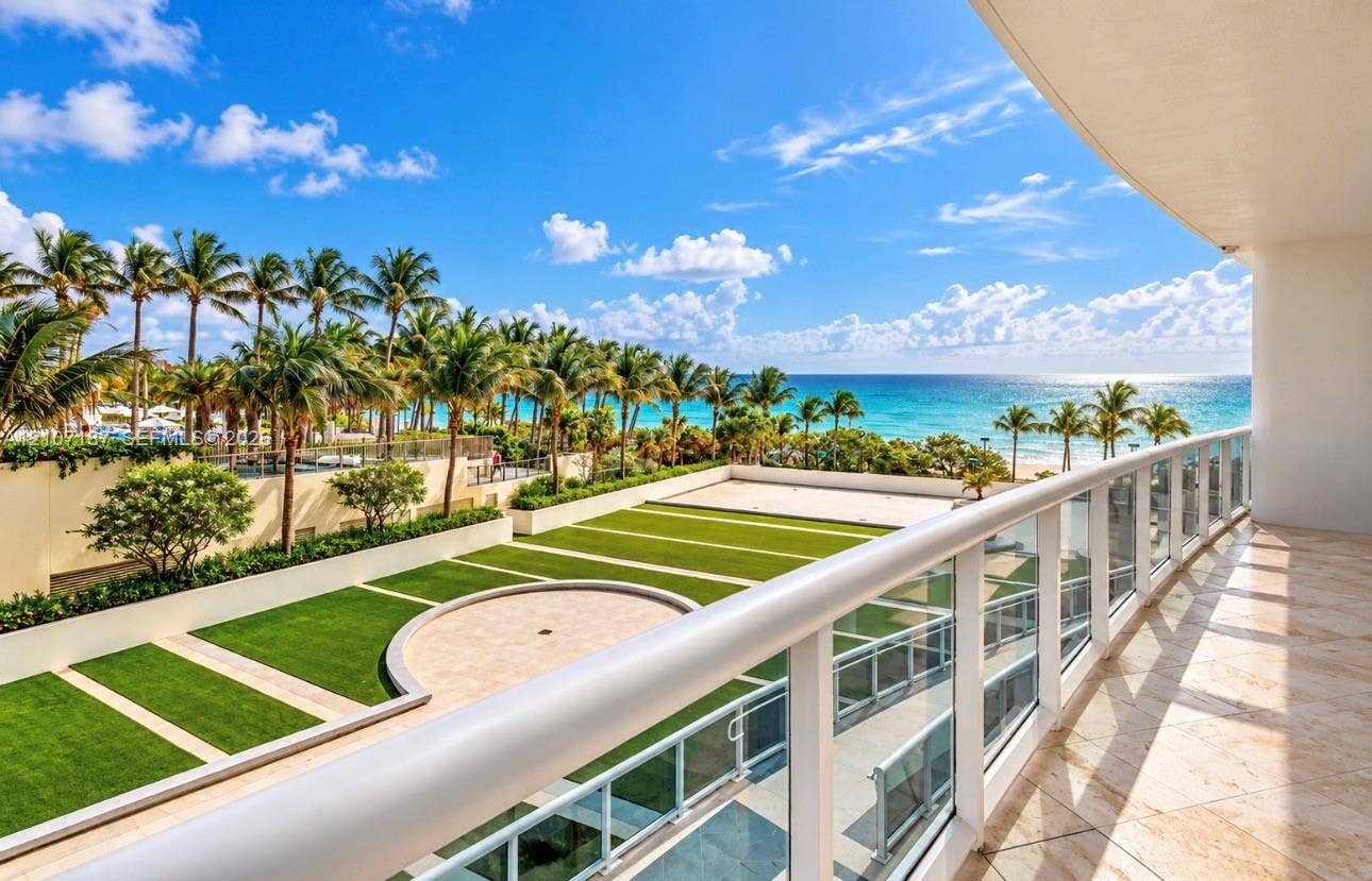 9601 Collins Avenue, #403, Bal Harbour, FL 33154 Private balcony featuring stone tile flooring and a glass-paneled railing overlooking a landscaped courtyard with artificial turf, stone accents, and a coastal backdrop