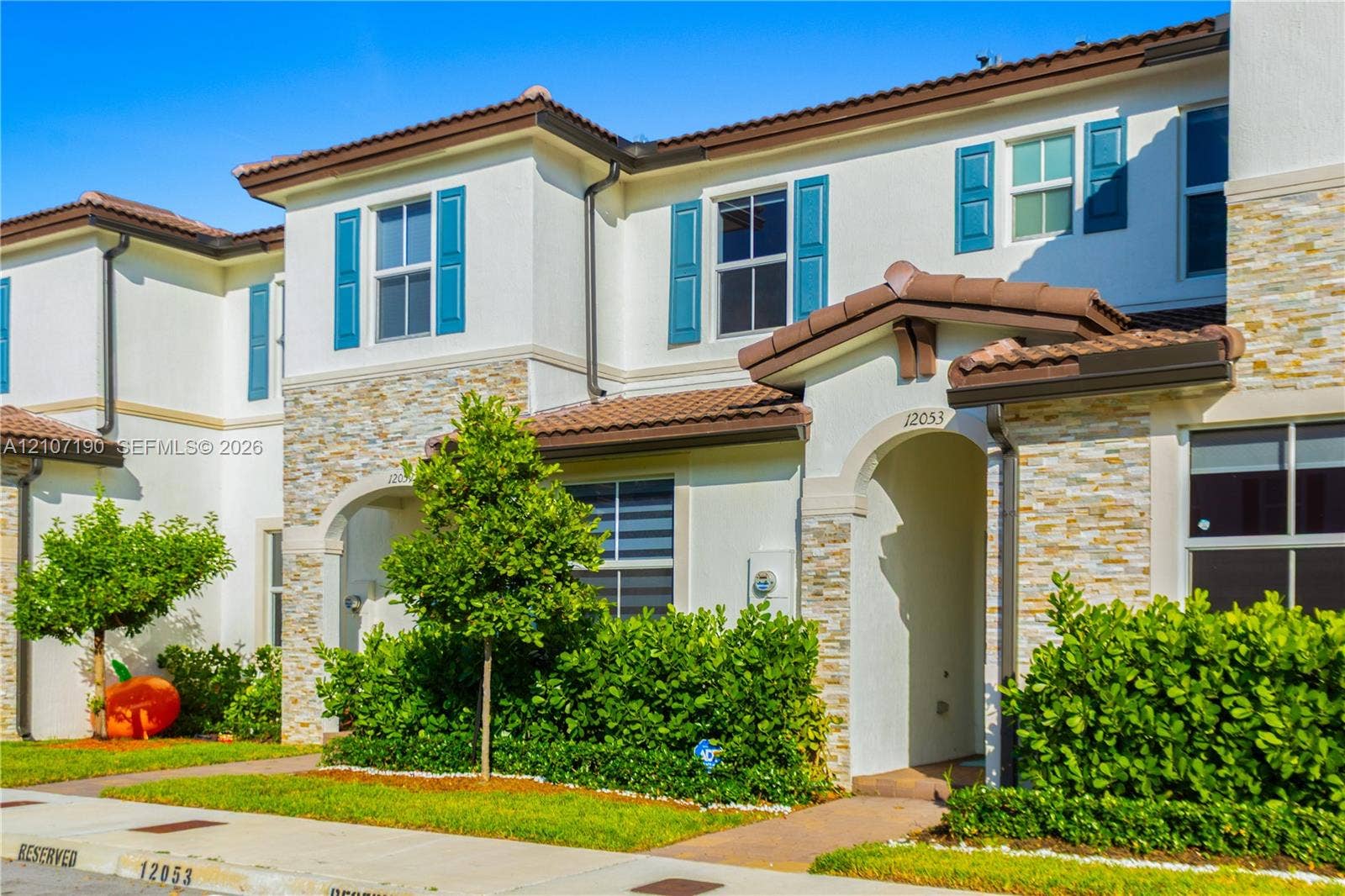 Two-story exterior featuring light-toned stucco walls, stacked stone veneer accents, blue louvered shutters, and a multi-gabled tile roof