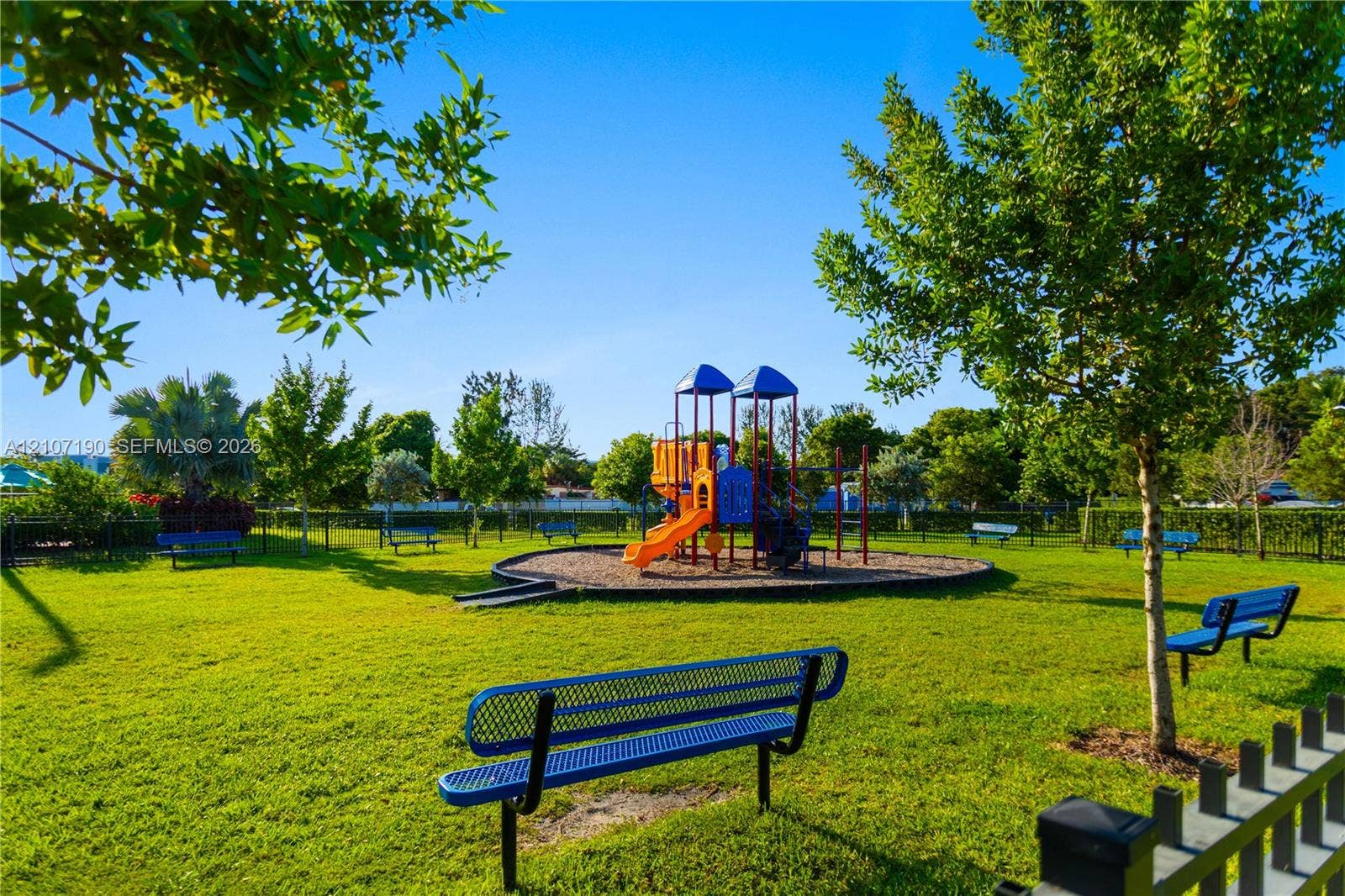 Outdoor recreational area featuring a playground structure with slides, a wood-mulch base, multiple metal benches, and established trees within a fenced perimeter