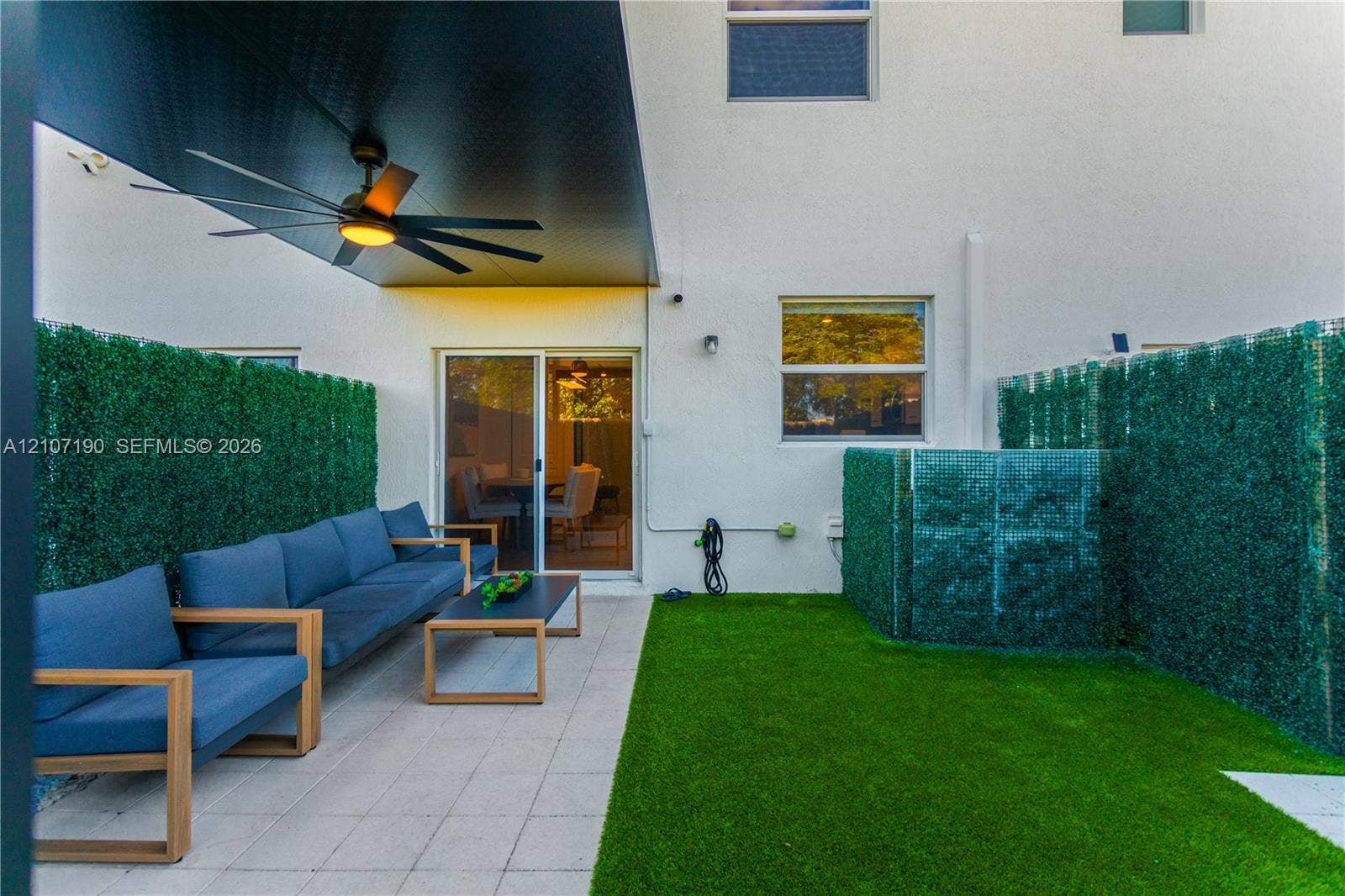 Covered patio featuring a high-contrast dark ceiling, integrated ceiling fan, light-toned concrete pavers, synthetic turf lawn, and perimeter privacy screening