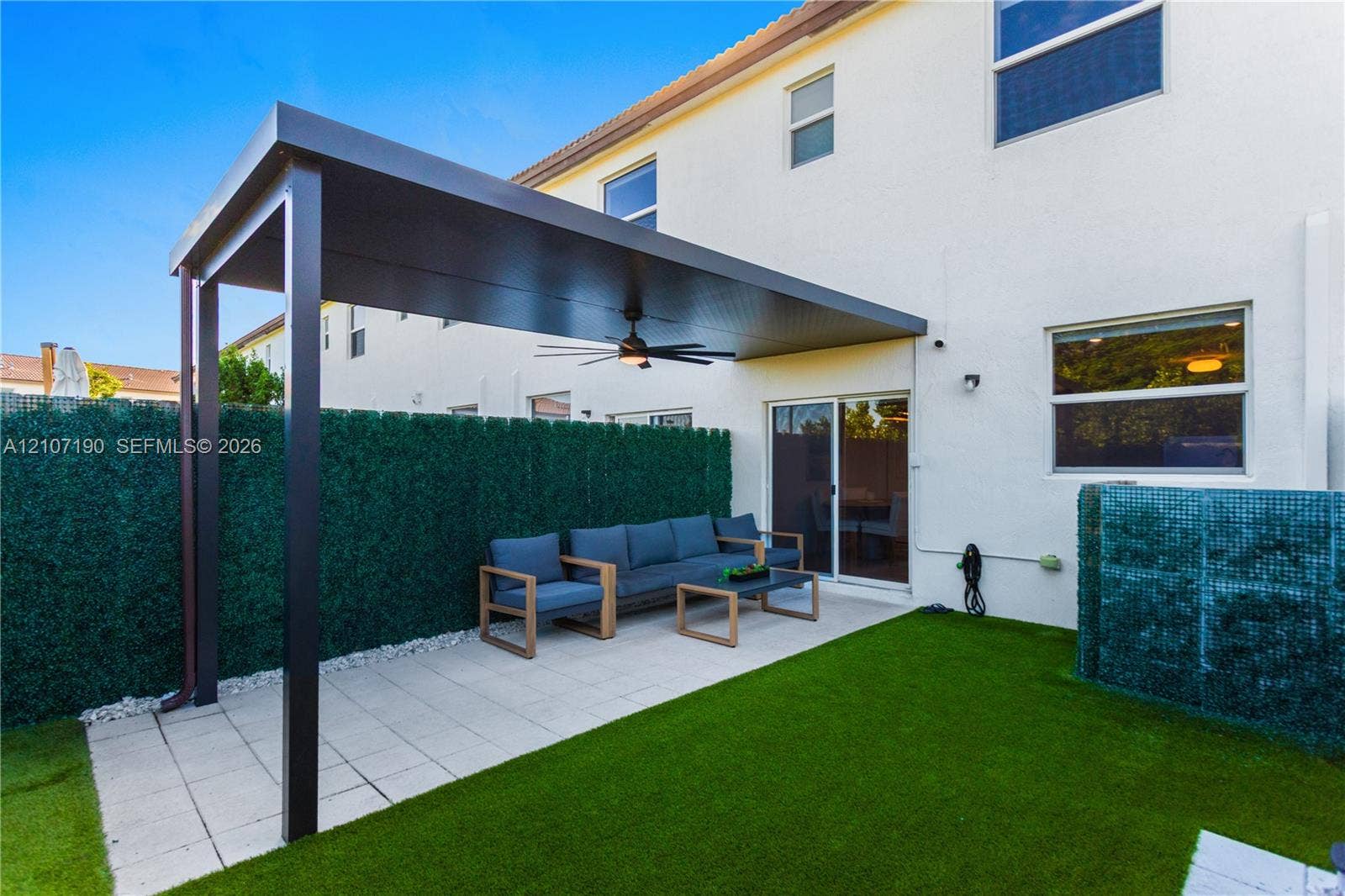 Rear patio featuring a metal-frame pergola with an integrated ceiling fan, paved flooring, synthetic lawn, and privacy screening