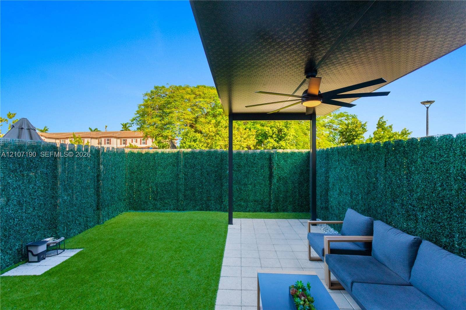 Covered patio featuring a metal roof, ceiling fan, and tiled flooring, accompanied by a synthetic turf lawn enclosed by faux-hedging privacy fencing