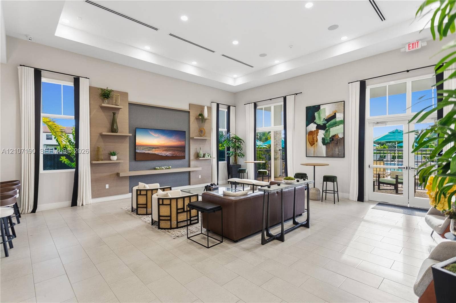 Spacious common area featuring recessed tray ceiling lighting, large-format tile flooring, a custom media wall with built-in shelving, and floor-to-ceiling glass-paned doors