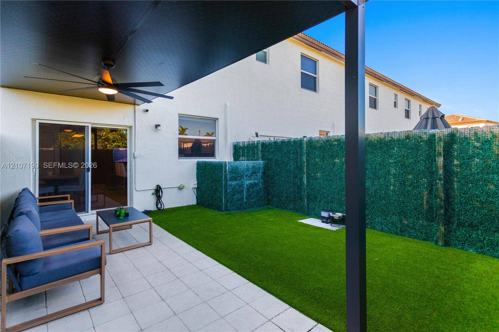 Covered patio with a mounted ceiling fan, tile flooring, and synthetic turf lawn enclosed by privacy screening