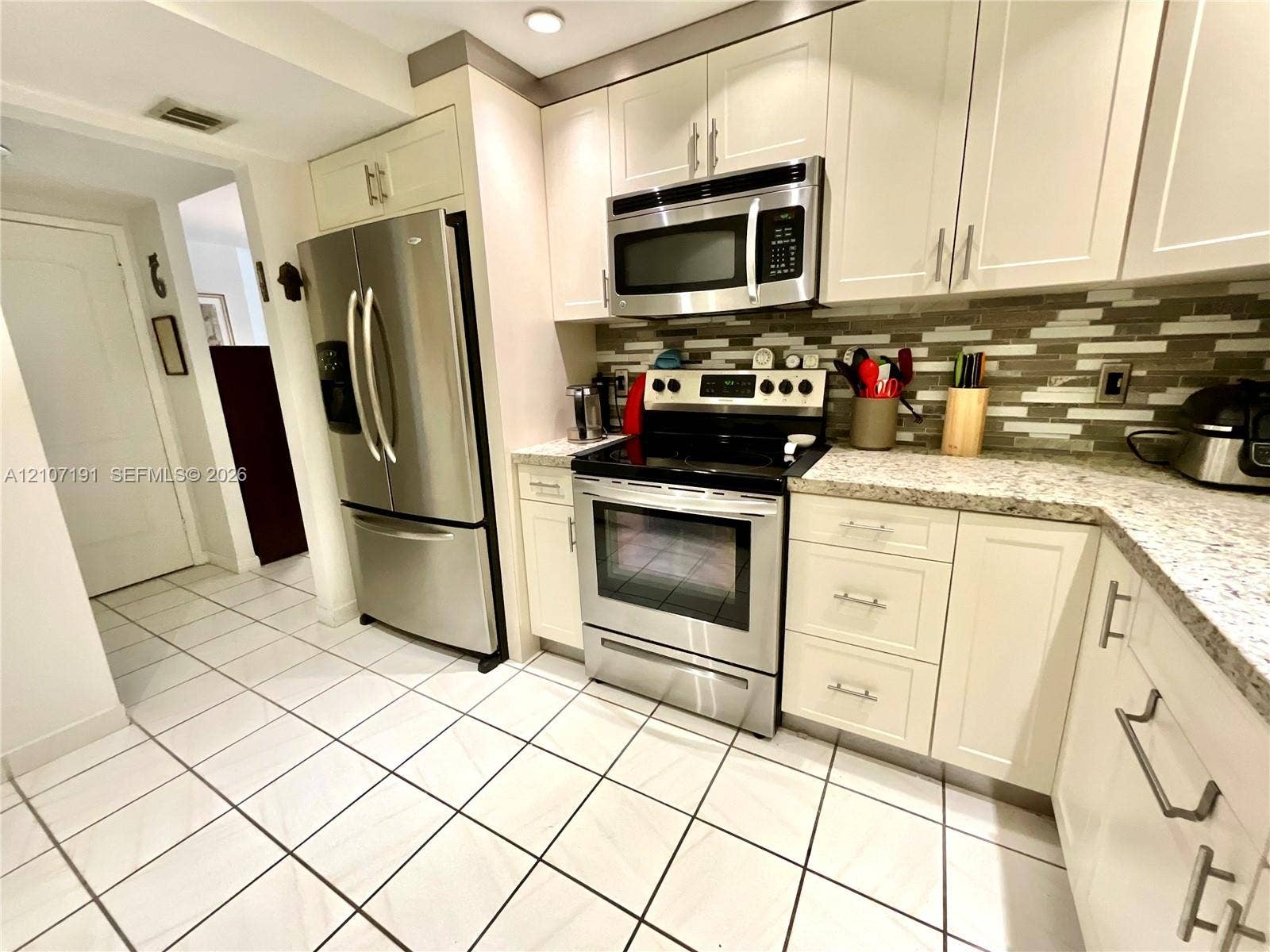 Kitchen featuring shaker-style cabinetry, stainless steel appliances, multi-tonal linear tile backsplash, patterned stone countertops, and tile flooring