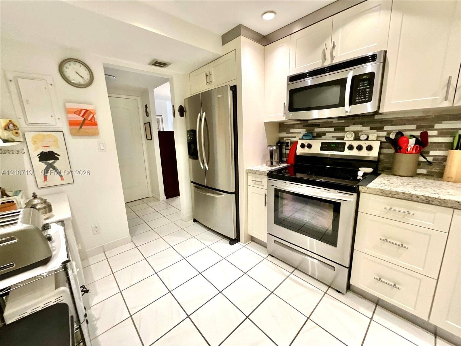 Kitchen featuring white shaker-style cabinetry, stainless steel appliances, multi-toned tile backsplash, stone-finish countertops, and ceramic tile flooring