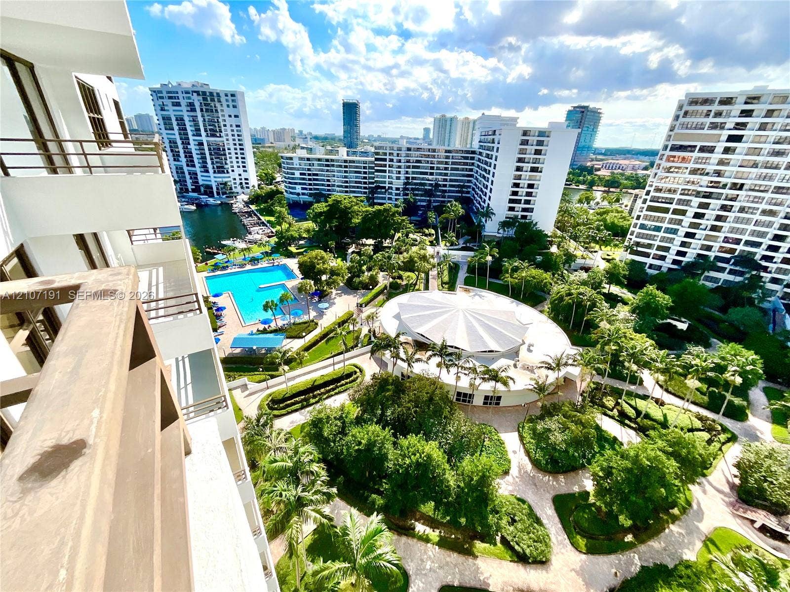Covered balcony with metal railings overlooking a community swimming pool, circular clubhouse, manicured landscaping, and mature palm trees