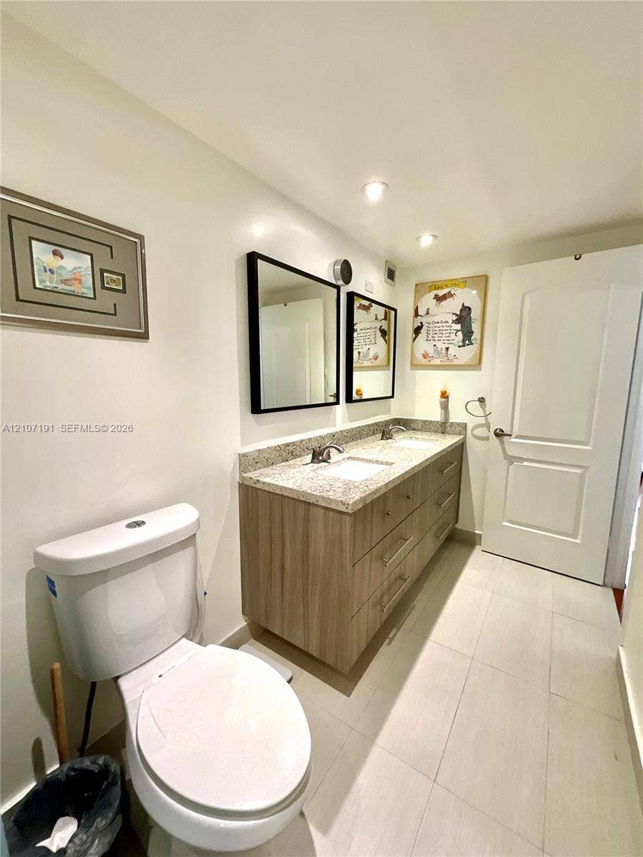Double vanity featuring light wood-finish cabinetry, stone-composite countertop with integrated dual sinks, two rectangular mirrors with black frames, and tile flooring